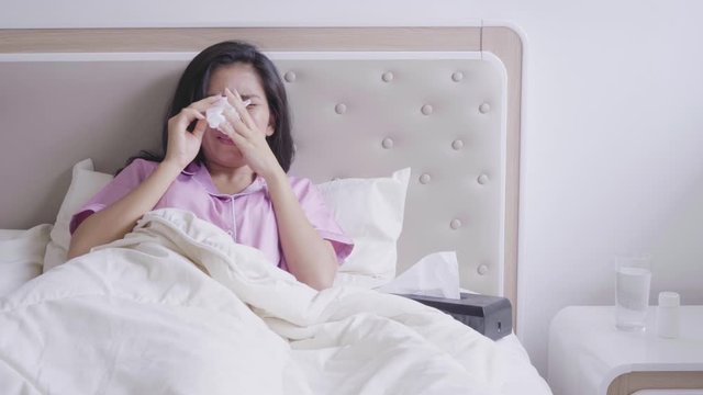 Sick young woman lying on bed under blanket and sneezing her nose into tissue. Shot in 4k resolution