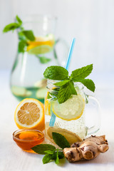 Healthy and tasty drink: homemade lemonade with lemon, mint, ginger and honey in transparent glass mug with blue straw. Small bowl of natural honey on the front. White background, copy space for text