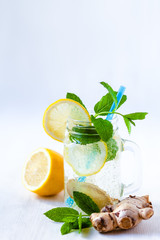 Healthy and tasty drink: homemade lemonade with lemon, mint, ginger in transparent glass mug with blue straw. Healthcare, concious lifestyle, detox. White background, jug in the background. Copy space