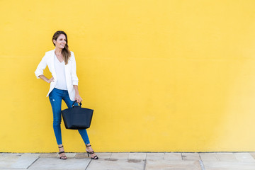 Smiling woman standing at a yellow wall holding a handbag - obrazy, fototapety, plakaty