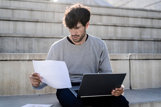 Man sitting on outdoor stairs using laptop and reviewing paper