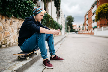 Spain, Torredembarra, smiling young man sitting on his skateboard at curb listening music with headphones