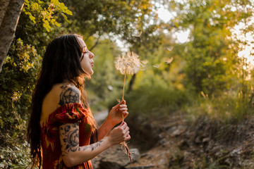 Young woman blows a big dandelion in a forest at sunset