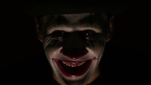 Scary clown looks at the camera and laughs terribly. A scary man in a clown makeup looks at the camera and laughs. Scary clown grimaces looking into camera .Halloween.