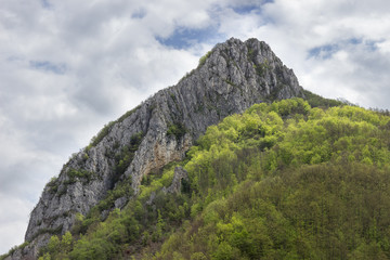 Impressive, rocky, pointy summit Asenovo kale, dramatic cloudy sky and beautiful sunlit, vivid green forest during early spring