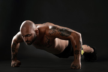 Athletic bald, tattooed man in black shorts and sneakers is posing against a black background. Close-up portrait.
