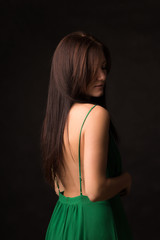 A beautiful long-haired brunette in a green dress with a bare back stands on a dark background. Romantic, pensive and sexy.