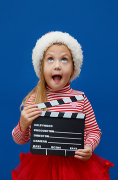 Smiling little child girl in Santa hat with a clapperboard on blue background. Christmas and New Year holiday concept