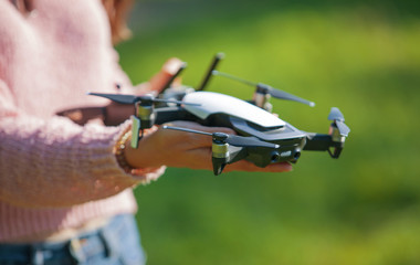 A young woman in a clearing in a park launches, checks, holds a drone, in one hand, in the other control panel.