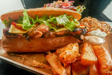 Ciabatta Grilled Chicken Sandwich  with Lettuce Cheese and Tomato on Wood Plate With Potato Wedges and a Salad Bowl with Mayonnaise Sauce and  Spicy Sauce and Fried Onions 