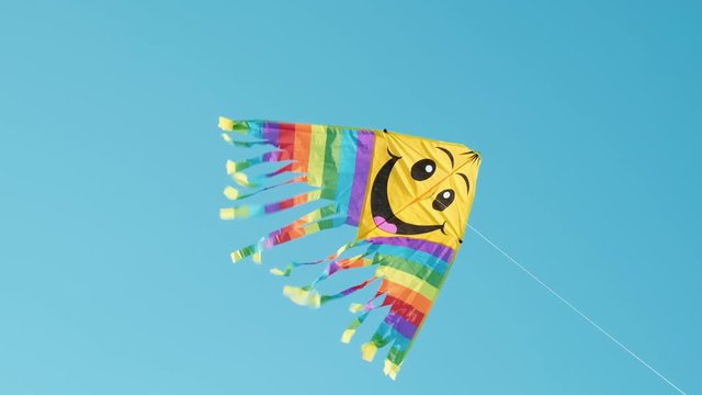 A colored kite with a smiley emoji hangs in the air against a blue sky on a sunny summer day at a city kite festival. The tail of a kite sways in the wind. Children's holiday. Lifestyle