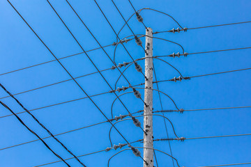 A low angle view of a tall wooden utility pole supporting overhead electric voltage cables with insulators, crossing t-junction lines against a bright blue sky.
