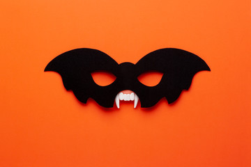 Conceptual halloween flat lay on a bright orange background. Black bat-mask and vampire fangs as a symbol of halloween parties.