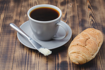 Coffee break with croissant butter knife and cup of black coffee on a dark wooden table.