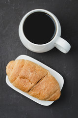 White mug with black coffee and croissant on dark stone table. Top view. Coffee break.
