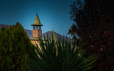 An abstract artistic cityscape of a clock tower's dome with defocused foreground of green vegetation and bushes, mountain range in the background, and clear blue sky above