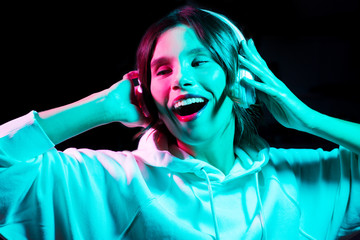 nightlife, technology and people concept - happy young woman in headphones wearing hoodie listening to music and dancing in neon lights over black background