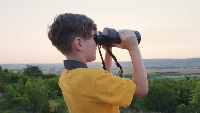 Happy boy looking through big binoculars beautiful landscape of green hills at sunset on a sunny day in summer and joyfully says something and shows his hand. Lifestyle