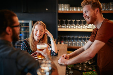 Cheerful friends leaning on bar counter, drinking beer and chatting with bartender. Night out.