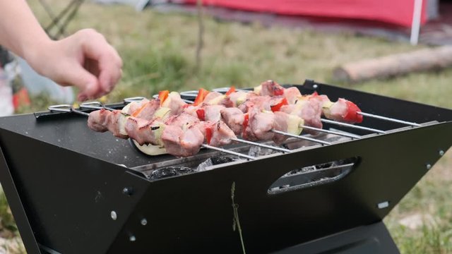 Family summer vacation camping with a tent. Woman's hand overturns a kebab barbecue on a small brazier against the background of a red tent in a clearing with green grass. Lifestyle