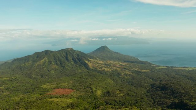 Tropical landscape: Mountains with forest, against blue sea with blue sky with clouds, top view, Camiguin, Philippines. Mountain landscape on tropical island with mountain peaks covered with forest