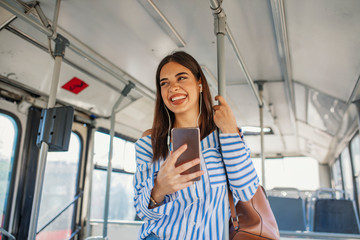 Young woman passenger standing with headphones and smartphone while moving in the modern tram,...