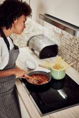 Mixed race woman in apron standing next to stove and stirring tomato sauce. On stove are saucepan...