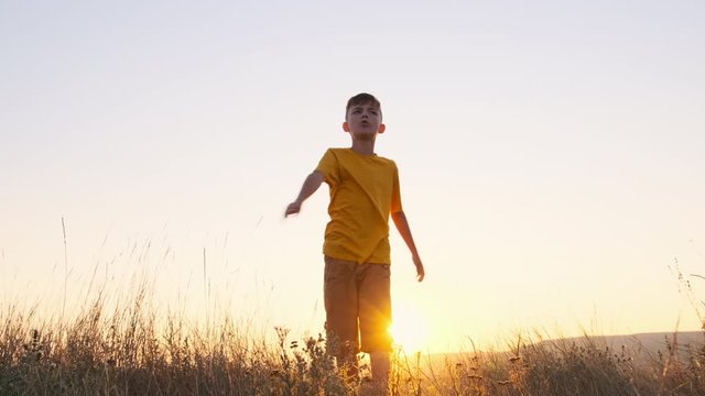 Slow motion happy boy in an orange T-shirt on summer meadow against the background bright sunset holds a white plane in his hand and smoothly turns and throws it up. Child plays. Lifestyle. Lens flare