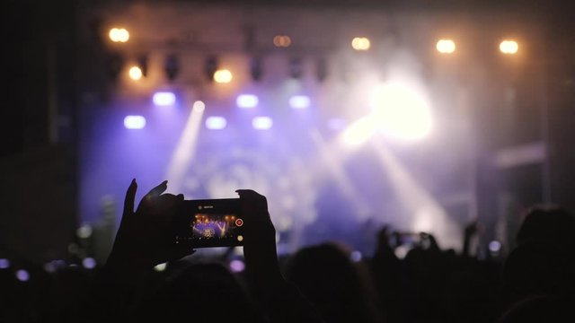 Rock concert Silhouette young woman is shooting smartphone. Musical rock festival. Laser stage show. Crowd of fans, rockers raised their hands, applause. Rock symbol made of finger. Lifestyle, freedom