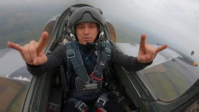 Caucasian pretty man in a helmet – navigator of pilot. Male relax and makes gestures goat or rocker. A gray combat fighter plane flying over field and river. Inside view from cockpit close up.