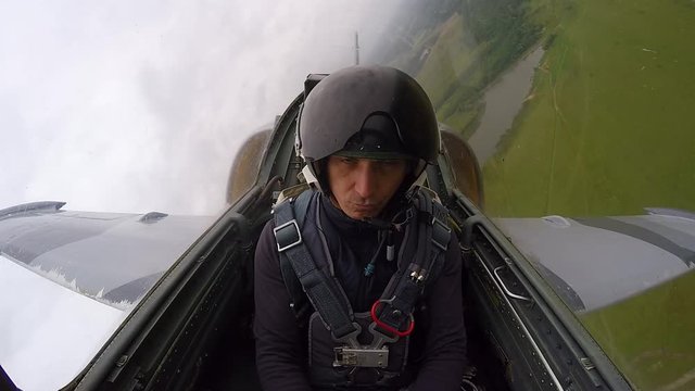 Caucasian man in a helmet - pilot of an gray training fighter plane flying and control yourself on the first real flight over green field in cloudy overcast sky. Inside view from cockpit close up.