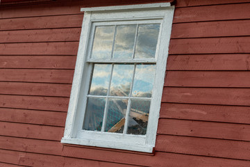 The exterior of an old red wooden shed wall with a white framed glass window. In the window is a reflection of the sky and you can see lobster pots stacked inside the building.
