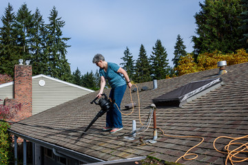 Rooftop view of suburban home, mature woman with leaf blower cleaning roof and gutter
