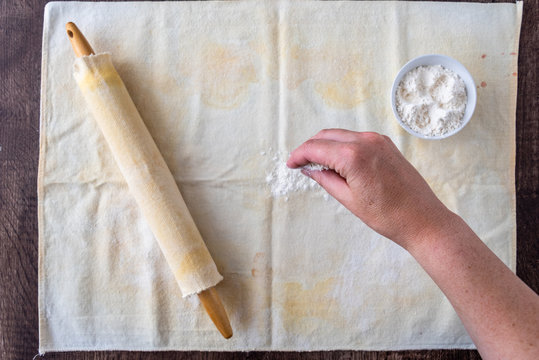 Woman’s hand dropping flour on pastry cloth, wood rolling pin with cloth cover, small bowl of flour