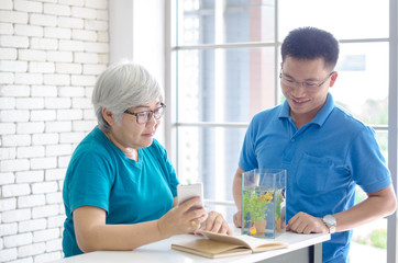 Fototapeta na wymiar Happy Asian senior woman reading a book and consult talking with her son with Gold fish in aquarium on white table, Happy day and good time family relationships concept