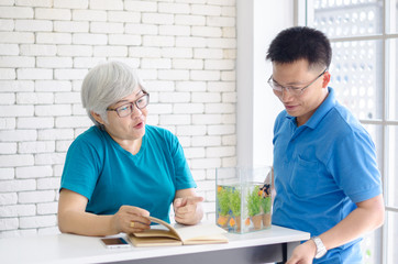 Happy Asian senior woman reading a book and consult talking with her son with Gold fish in aquarium on white table, Happy day and good time family relationships concept
