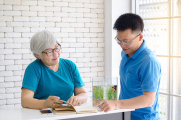 Happy Asian senior woman reading a book and consult talking with her son with Gold fish in aquarium on white table, Happy day and good time family relationships concept