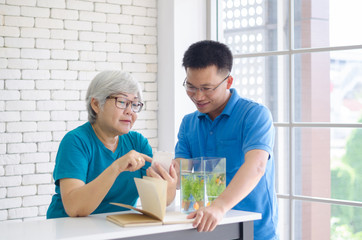 Happy Asian senior woman learning to use smartphone teach by her son with Gold fish in aquarium on white table, Happy day and good time family relationships concept