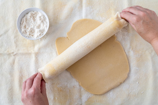 Woman’s hands on wood rolling pin, cloth covered, rolling out cookie dough on pastry cloth, small bowl of flour
