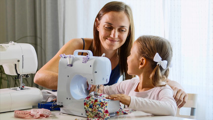 A girl is using a small sewing machine, a woman using a needle. A mother and a daughter are at home together, day time. Mommy explains a little child how to work with equipment and fabric. Close-up.