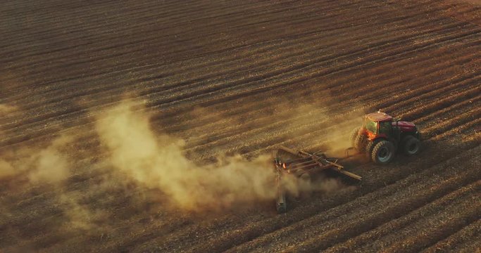 Aerial view of a red tractor farming at sunset kicking up golden dusty soil into the air, climate change apocalypse fallow fields