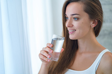 Happy attractive joyful healthy drinking woman holding glass of clear purified water in the morning in the kitchen at home. Healthy lifestyle