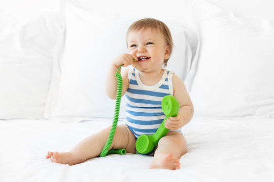 Laughing baby sitting on bed chewing on green phone