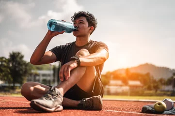 Fotobehang De jonge man droeg alle delen van zijn lichaam en dronk water om zich voor te bereiden op het joggen op de atletiekbaan rond het voetbalveld. © Day Of Victory Stu.