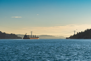 Obraz premium A large cargo ship - bulk-carrier, sailing along the blue waves of the strait, in the light of the evening sun, is approaching a bridge over the Bosphorus Strait.