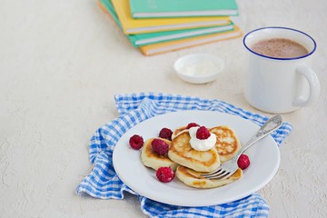 Breakfast, pancakes with sour cream and raspberries on a white plate on a light concrete background. Child's breakfast in front of the school. School breakfast concept.