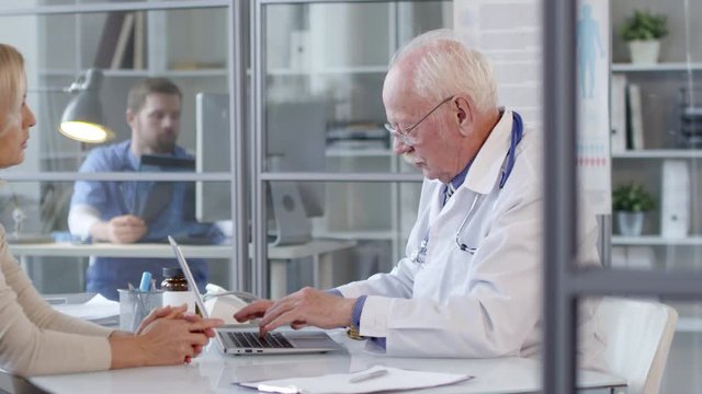 Experienced senior doctor typing on laptop and talking with female patient while male physician examining x-ray image behind glass wall in the background
