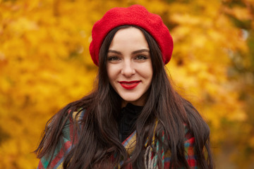Colorful, cozy autumn, fashion concept. Outdoor portrait of young beautiful smiling girl wearing red beret. Model wrapped in checkered blanket, looking smiling directly at camera, expressing happyness