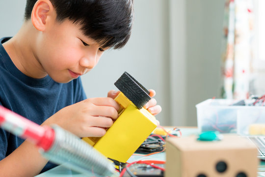Closeup: Smart looking Asian boy working with circuits, wires, computer chip, OLED, motor, wheels on his robotics project. Science, Technology, Engineering and Mathematics (STEM) education concept.