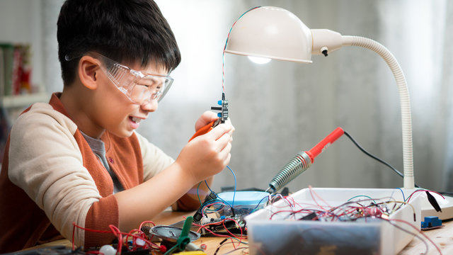 Smart looking preteen / teenage Asian boy using plier to repair, fix solid state relay on desk, smiling, wearing protective / safety glasses with concentration. Electronics and Science school project.